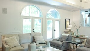 Un salón amplio con un sofá de color claro, sillones y una mesa de centro. Dos grandes ventanales permiten la entrada de luz natural.