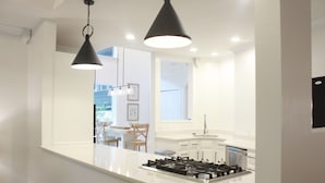 Una cocina moderna con una isla central, muebles blancos y una estufa a gas. Hay lámparas colgantes sobre la isla e iluminación empotrada en el techo.