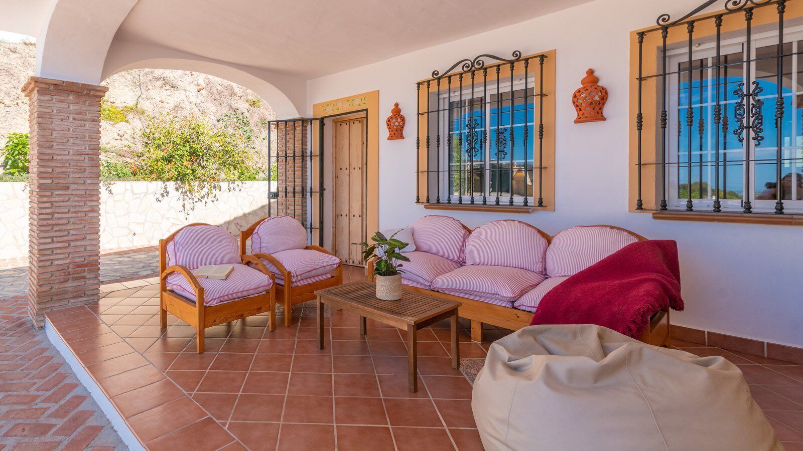 A covered patio with a brick column, two wooden chairs, a sofa, a table, and a bean bag.