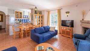 Een woonkamer met een blauwe bank, een televisie en een open haard. Er is een keukenhoek met een magnetron en een eettafel met stoelen.