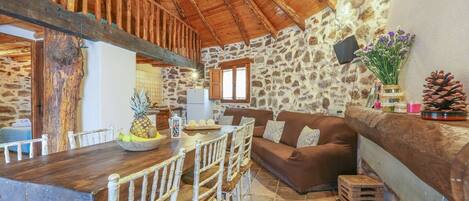 A rustic interior with a stone wall, wooden beams, a dining area with a wooden table and chairs, a brown sofa, and a kitchen in the background.