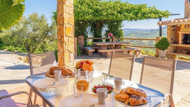 Un espacio para comer al aire libre con pilares de piedra, una mesa dispuesta con pan, fruta y bebidas, y vistas al campo.