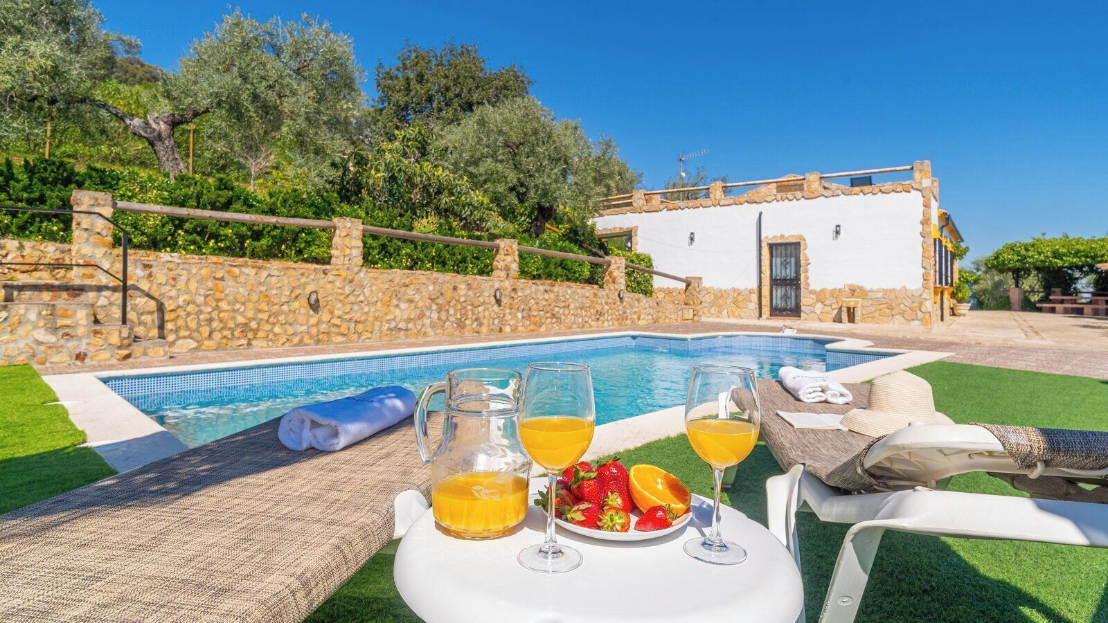 Una mesa junto a la piscina con bebidas y fruta, un muro de piedra y un edificio con tejado de azulejos.