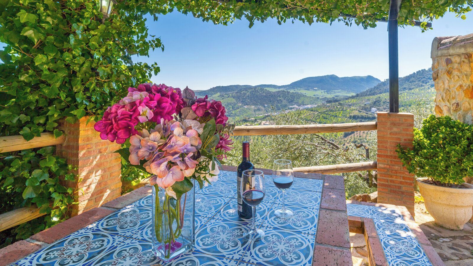 A table with a bottle and glasses, surrounded by a blue patterned tablecloth, with a view of hills in the background.
