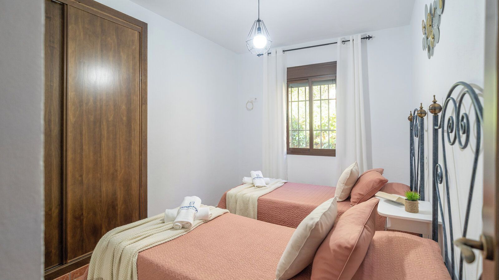 Une chambre avec deux lits, une armoire, une fenêtre avec des rideaux, une petite table avec une plante et un cadre de lit en métal.