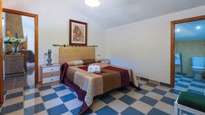 A hotel room with a checkered floor, a bed with a quilt, a bedside table with a lamp, a framed picture on the wall, and a bathroom visible through an open door. - Casa Camino del Río Pozo Alcón by Ruralidays (Jaén)