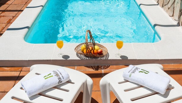 Zona de piscina con dos sillas de salón blancas, cada una con una toalla y un vaso de jugo de naranja. Una cesta de mimbre con frutas y un vaso de jugo de naranja se encuentra en el borde de la piscina.