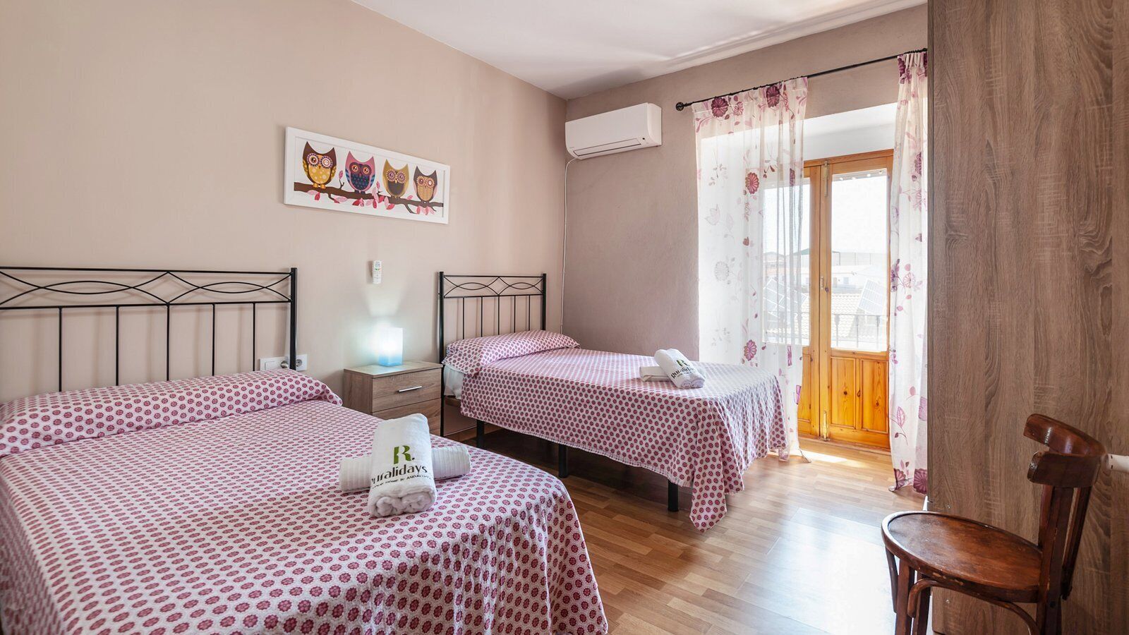 Une chambre d’hôtel avec deux lits, une chaise en bois, une table de chevet avec une lampe, un tableau représentant des hiboux et une fenêtre avec des rideaux.