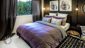 A bedroom with a large bed, dark curtains, a window with a view of greenery, two wall-mounted light fixtures, and a round mirror. - Indigo Boutique Apartment-2 km to city  (Wayville)
