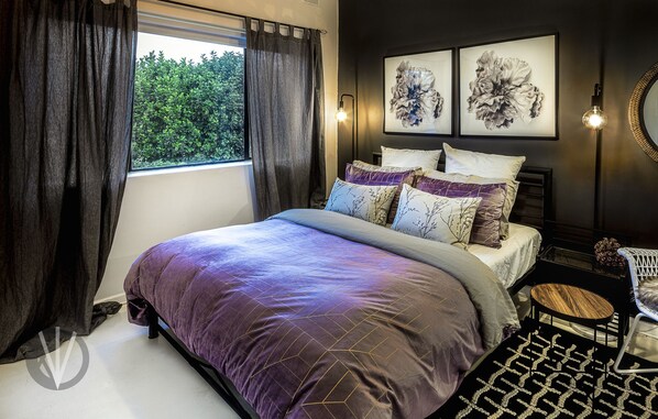 Une chambre avec un grand lit, des rideaux foncés, une fenêtre donnant sur un paysage verdoyant, deux appliques murales et un miroir rond.