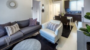 A modern living room with a grey sofa, a light blue armchair, and a dining area with a table and chairs. There is a kitchen visible in the background with cabinets and a window. - Indigo Boutique Apartment-2 km to city  (Wayville)