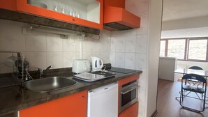 Een kleine keuken met oranje kastjes, een witte koelkast, een zwart aanrechtblad en een raam dat voor daglicht zorgt.