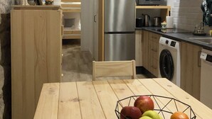 Een kleine keuken met een roestvrijstalen koelkast, een magnetron en een wasmachine. Een houten eettafel met een schaal fruit staat op de voorgrond.