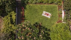 Eine Person liegt auf einer Decke in einem Garten mit rotem und weißem Zaun, umgeben von viel Grün und einer Steinmauer.