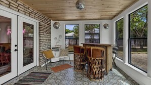 A cozy room with a stone wall, a wooden ceiling, and large windows. There is a white sofa, a small table, and two chairs. - Eclectic Fun Haus With Hot Tub - 3 Min Drive to Main (Fredericksburg)