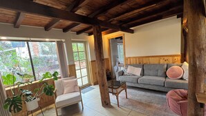 Un salon chaleureux avec un canapé gris, un fauteuil blanc et une table basse en bois. Une grande fenêtre, orn ée de rideaux, offre une vue sur la verdure environnante.