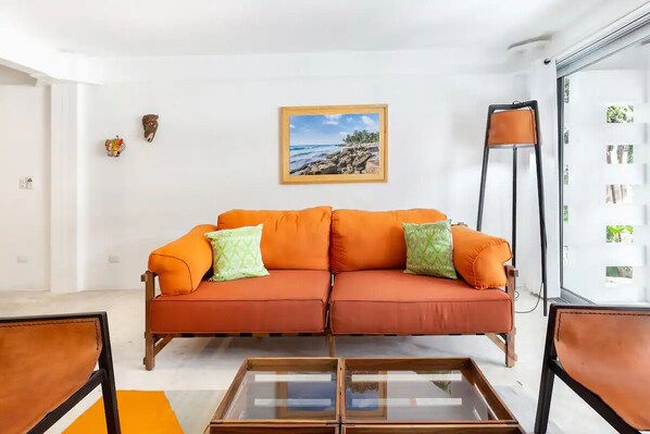 A living room with a sofa, two cushions, a glass coffee table, and a framed picture on the wall. - Design Apartment, 2 Bedroom, Deck and Garden View (Tulum)