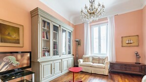 A living room with a television on a glass stand, a red table, a white sofa, a large cabinet, and a chandelier. - GuestHost - Elegant apartment of 80 sqm, able to accommodate up to 5 people, located on the 2nd floor of a building equipped with a lift (NOT suitable for disabled people - some steps to access). The property is located in the Stibbert/Statuto area, a li (Firenze)