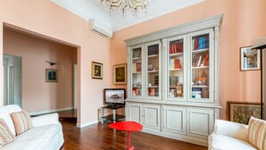 A living room with a white sofa, a red stool, a white cabinet with books, and a television. - GuestHost - Elegant apartment of 80 sqm, able to accommodate up to 5 people, located on the 2nd floor of a building equipped with a lift (NOT suitable for disabled people - some steps to access). The property is located in the Stibbert/Statuto area, a li (Firenze)