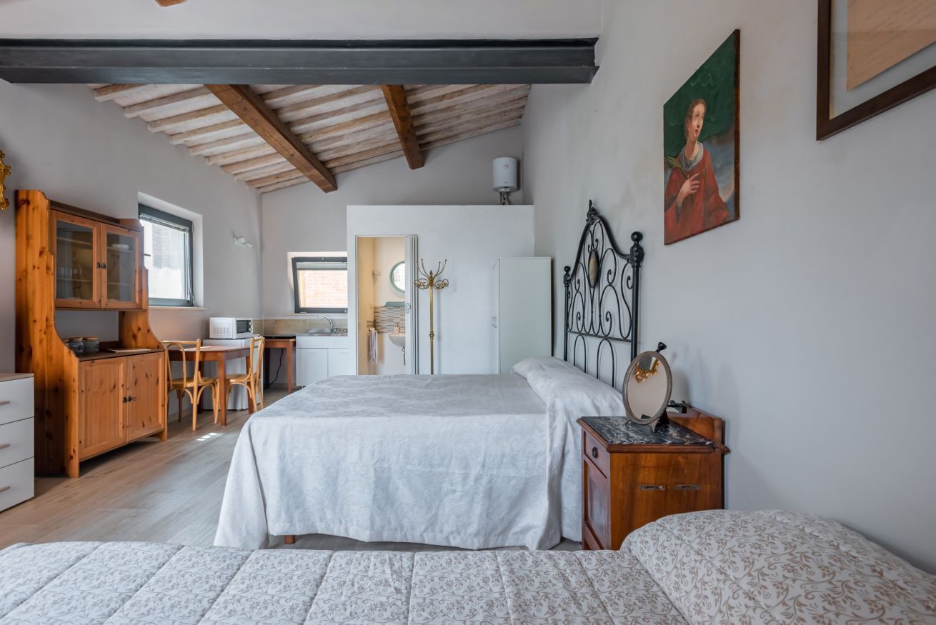 Camera da letto con un letto, un comodino e una cassettiera. Si vede una finestra e una porta che dà su un'altra stanza.