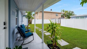 A covered patio with a rocking chair and a small table, a well-maintained lawn, and a white fence in the background.