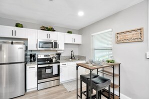 Una cocina moderna con armarios blancos, electrodomésticos de acero inoxidable, una mesa de madera con taburetes y un letrero en la pared que dice 'stay awhile'.