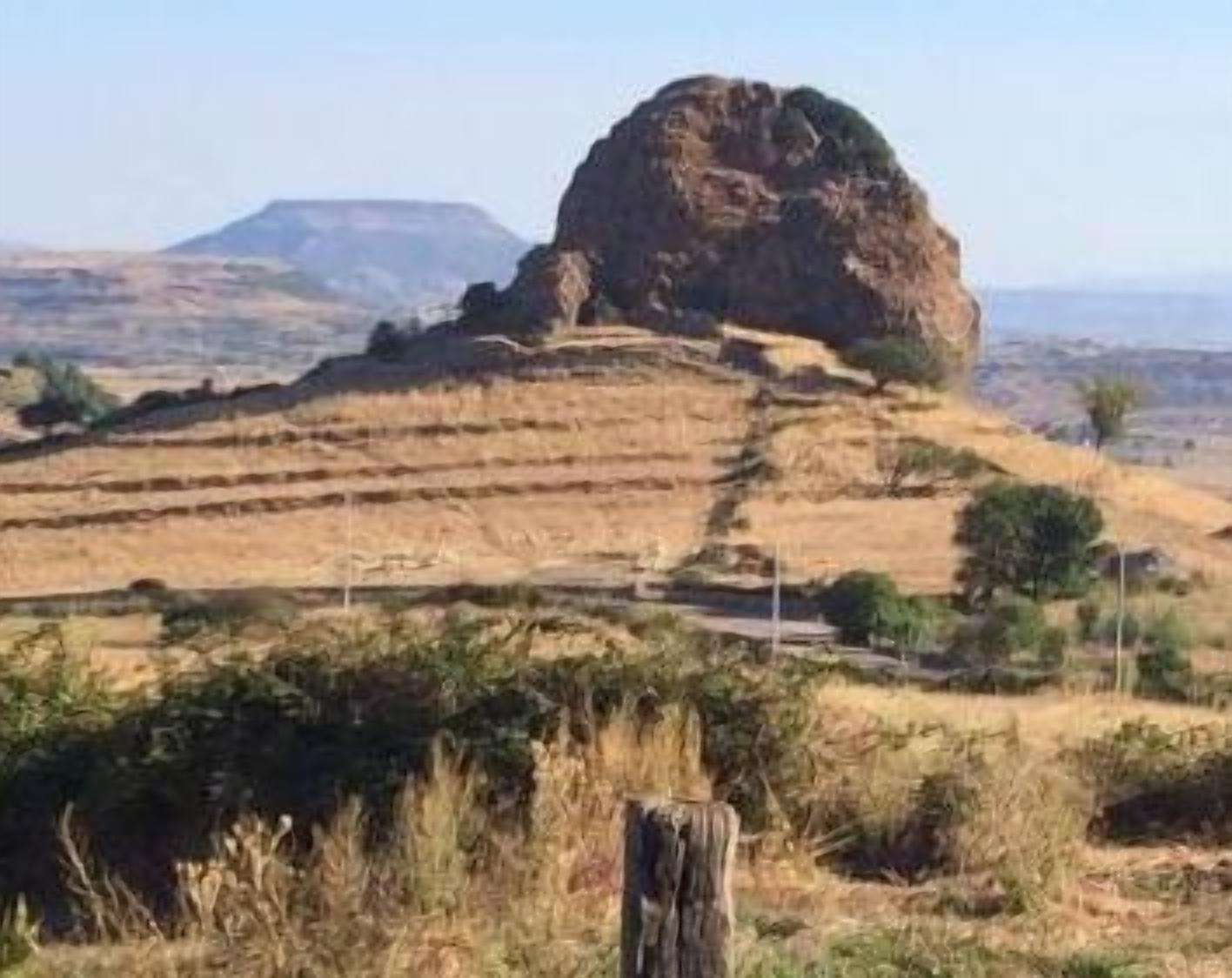 A large rock formation in a dry, hilly landscape with sparse vegetation.