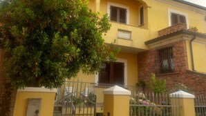 Um edifício amarelo de dois andares, com base de pedra, um portão de metal e uma árvore em primeiro plano.