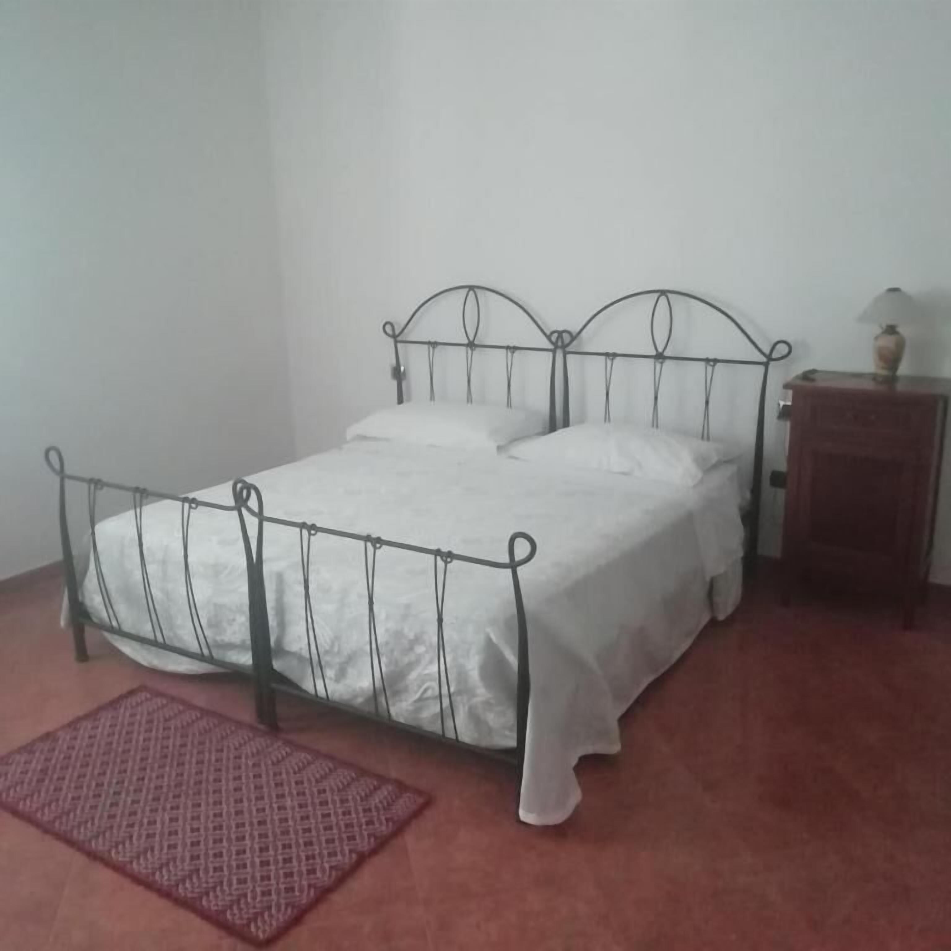 Una cama individual con estructura metálica, ropa de cama blanca y una mesita de noche con lámpara.