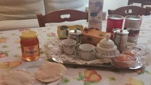 Uma mesa de jantar com toalha de mesa florida, um prato com pães achatados, um pote de mel, uma caixa de chá, um recipiente de leite e várias latas de café.