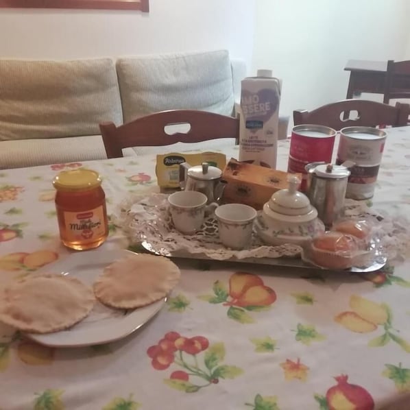 Una mesa de comedor con mantel floral, un plato de panes planos, un tarro de miel, una caja de té, un cartón de leche y varias latas de café.