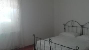Una cama individual con estructura metálica, una colcha blanca sencilla y un tapete rojo al pie de la cama.