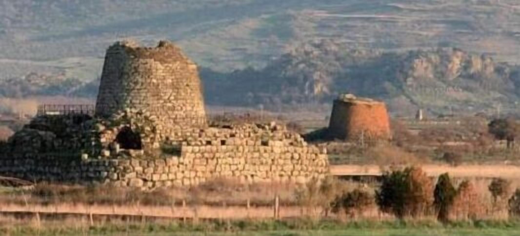 Una estructura histórica de piedra con una torre cilíndrica y un edificio rectangular, rodeada de campos abiertos y colinas distantes.