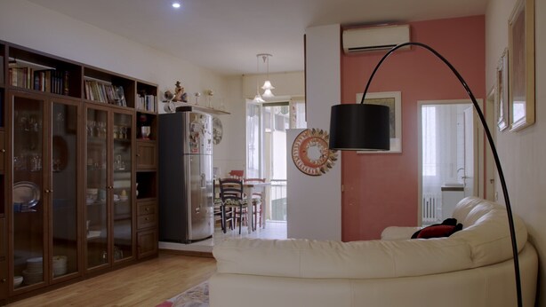Een woonkamer met een witte bank, een houten kast met glazen deuren, een eethoek met stoelen en een tafel, en een gang met een deur naar een andere kamer.