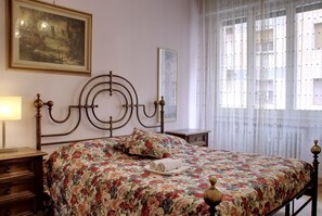 Une chambre à coucher avec une couvre-lit à motifs floraux, une tête de lit en bois, une table de chevet avec une lampe et une fenêtre ornée de rideaux légers.