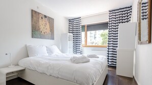A modern bedroom with a large bed, a bedside table, a radiator, and a window with striped curtains.
