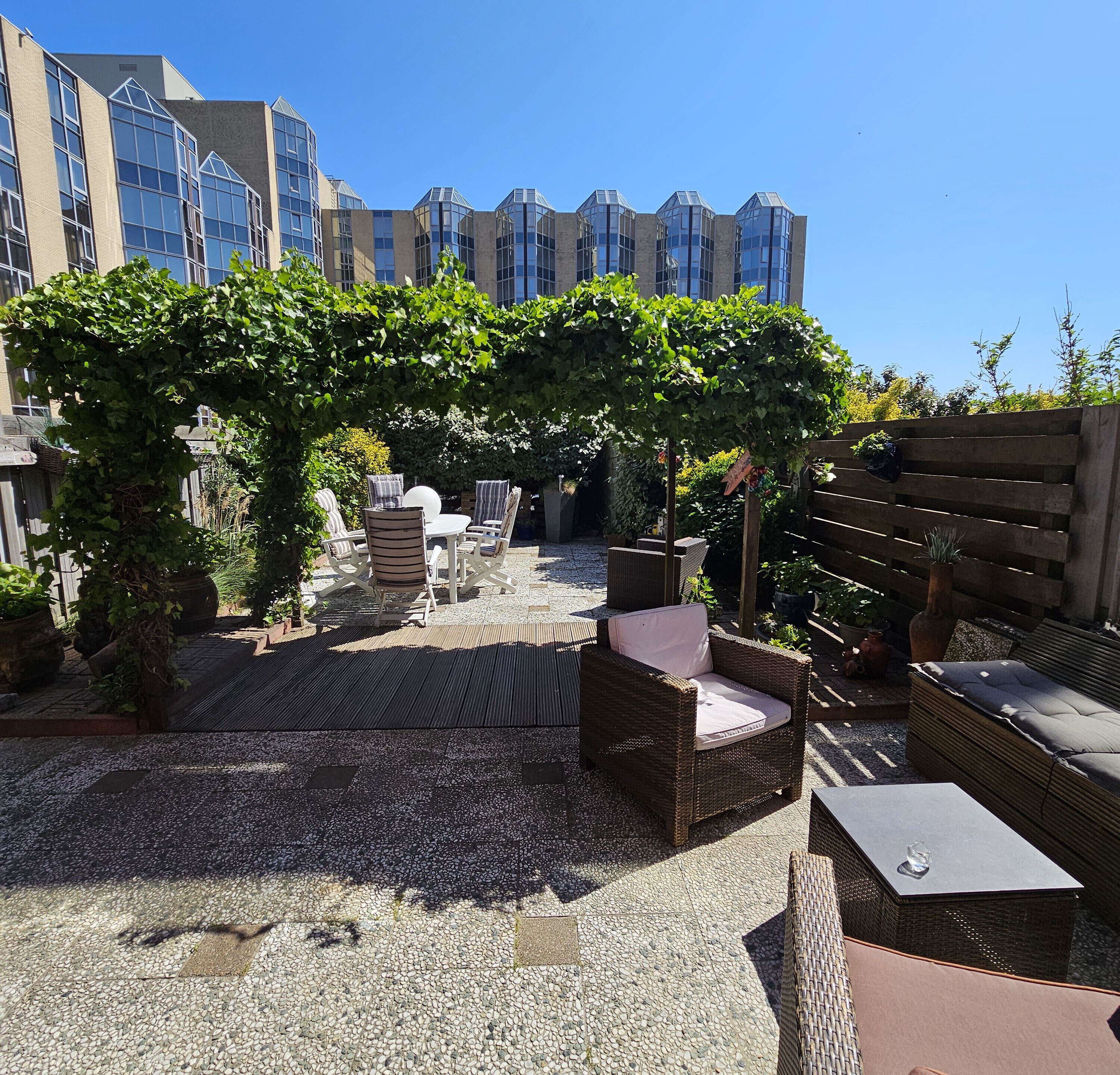 An outdoor seating area with wicker furniture, a pergola covered with greenery, and a view of modern buildings in the background.