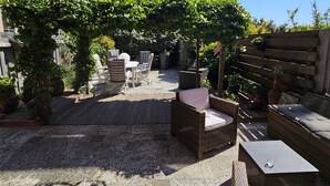 An outdoor seating area with wicker furniture, a pergola covered with greenery, and a view of modern buildings in the background.