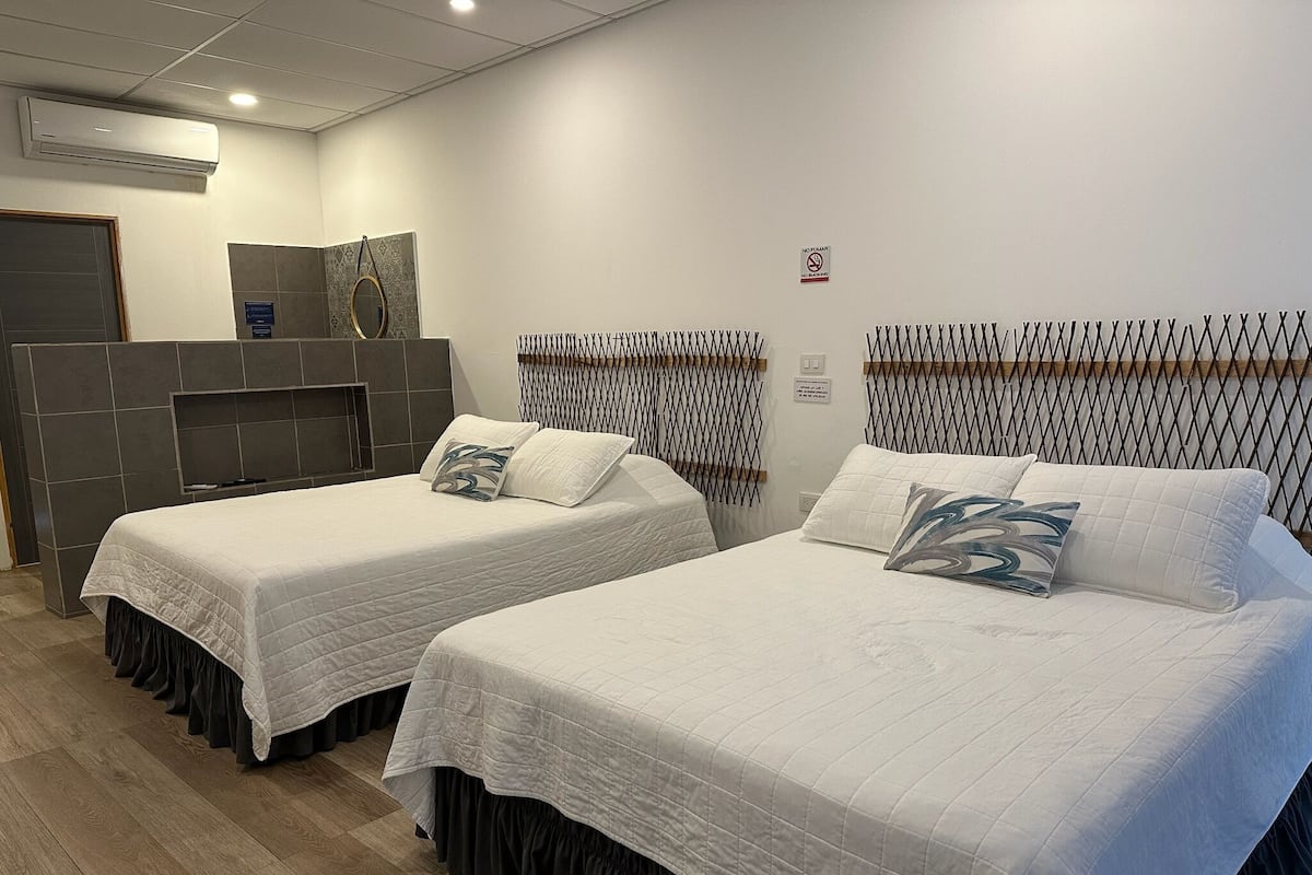 Two single beds with white bedding and dark skirts, a grey tiled wall, a grey cabinet, and a wall-mounted air conditioner.