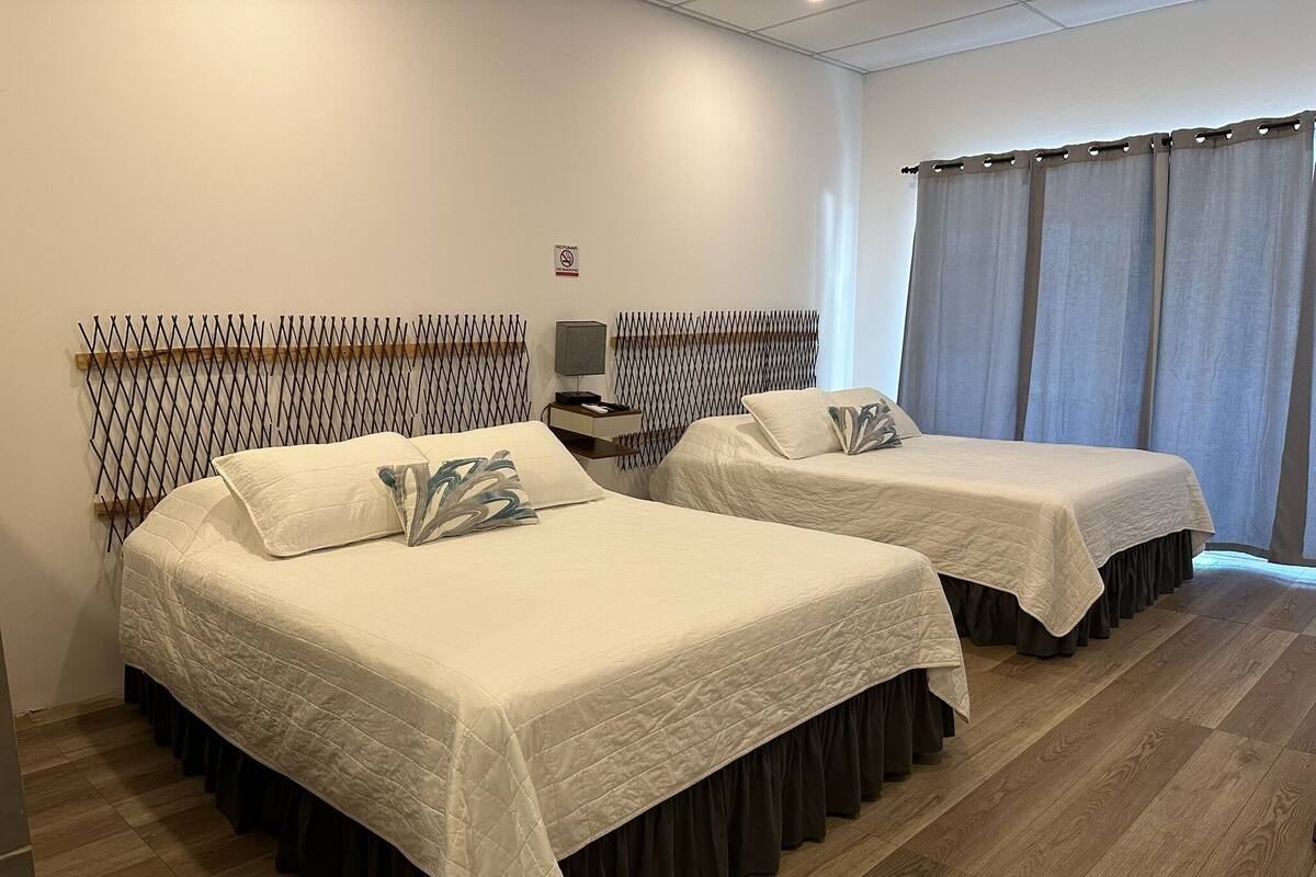 Two beds with white bedding and dark skirts, a wooden headboard, a nightstand with a lamp, and a window with curtains.