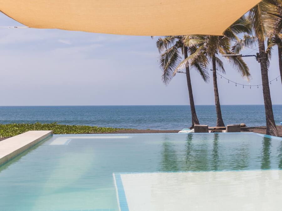 A pool with a view of the ocean, palm trees, and a shaded area.