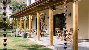 Un área de estar al aire libre con techo, con sillas y mesas de madera, rodeada de plantas en macetas y un camino de grava blanca.