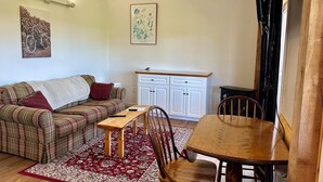 A living room with a plaid sofa, a wooden coffee table, and a dining table with chairs. There is a kitchen area with white cabinets and a countertop. - Waterfront Two Bedroom Newly Built Cottage, Beach, Covered Deck, Firepits  (New Glasgow)