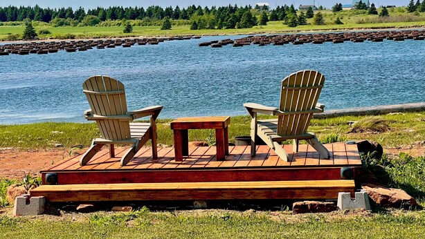 A wooden deck with two Adirondack chairs and a small table overlooking a body of water with a forested shoreline. - Waterfront Two Bedroom Newly Built Cottage, Beach, Covered Deck, Firepits  (New Glasgow)