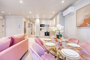 Ein modernes Wohnzimmer mit einem Esstisch aus Glas, gedeckt für sechs Personen, rosa Sofas und einer Küche im Hintergrund.
