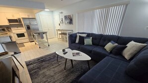 Un salon moderne avec un canapé bleu foncé, une table basse blanche et un coin cuisine à l’arrière-plan.