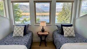 A room with two beds, a small table with a lamp, and large windows offering a view of a lake and mountains. - Columbia River Waterfront with Private Dock near Gamble Sands Golf Course! (Brewster)