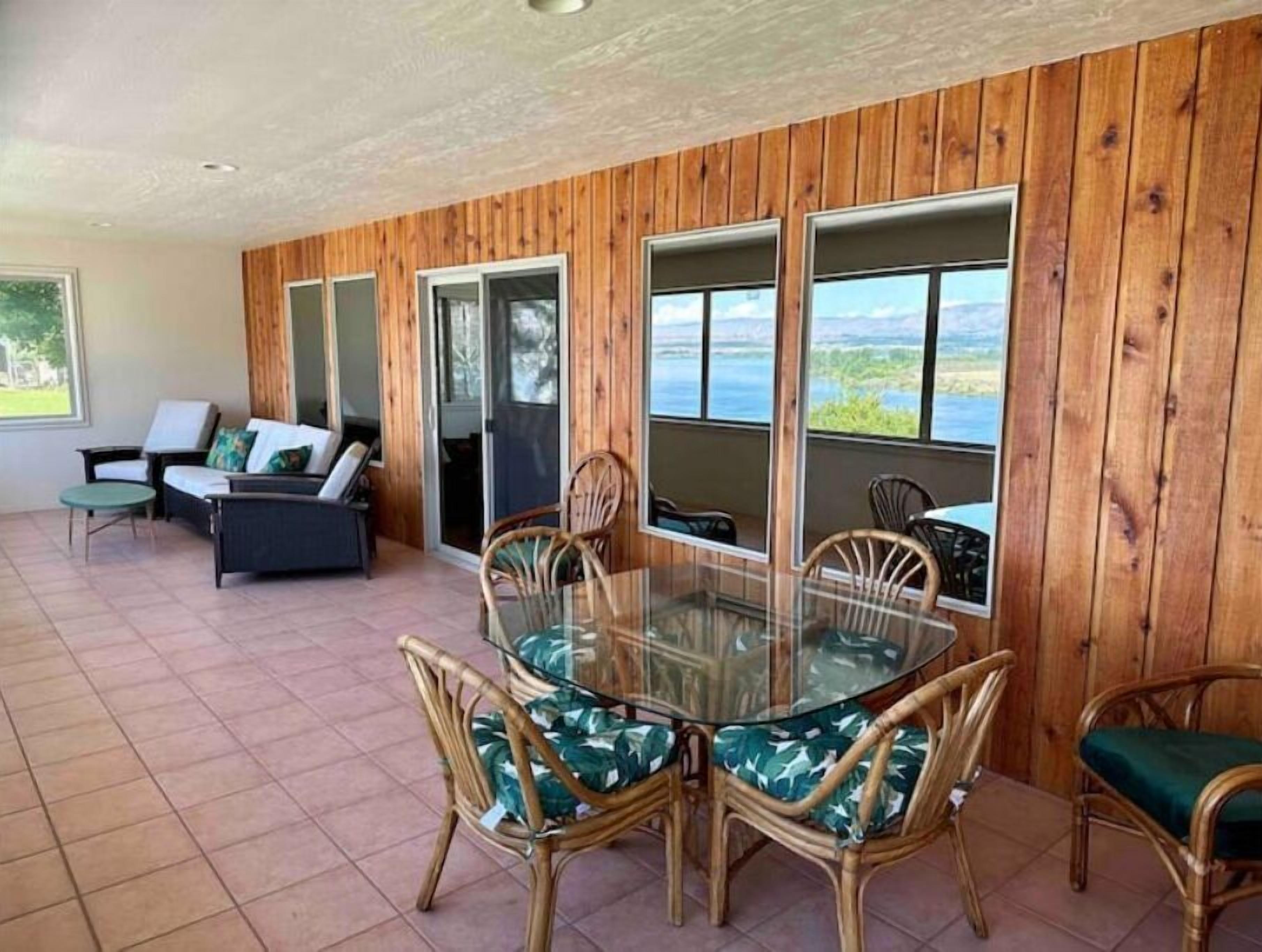 Uma sala de jantar com mesa de vidro e cadeiras de vime, paredes com painéis de madeira e vista para o exterior.