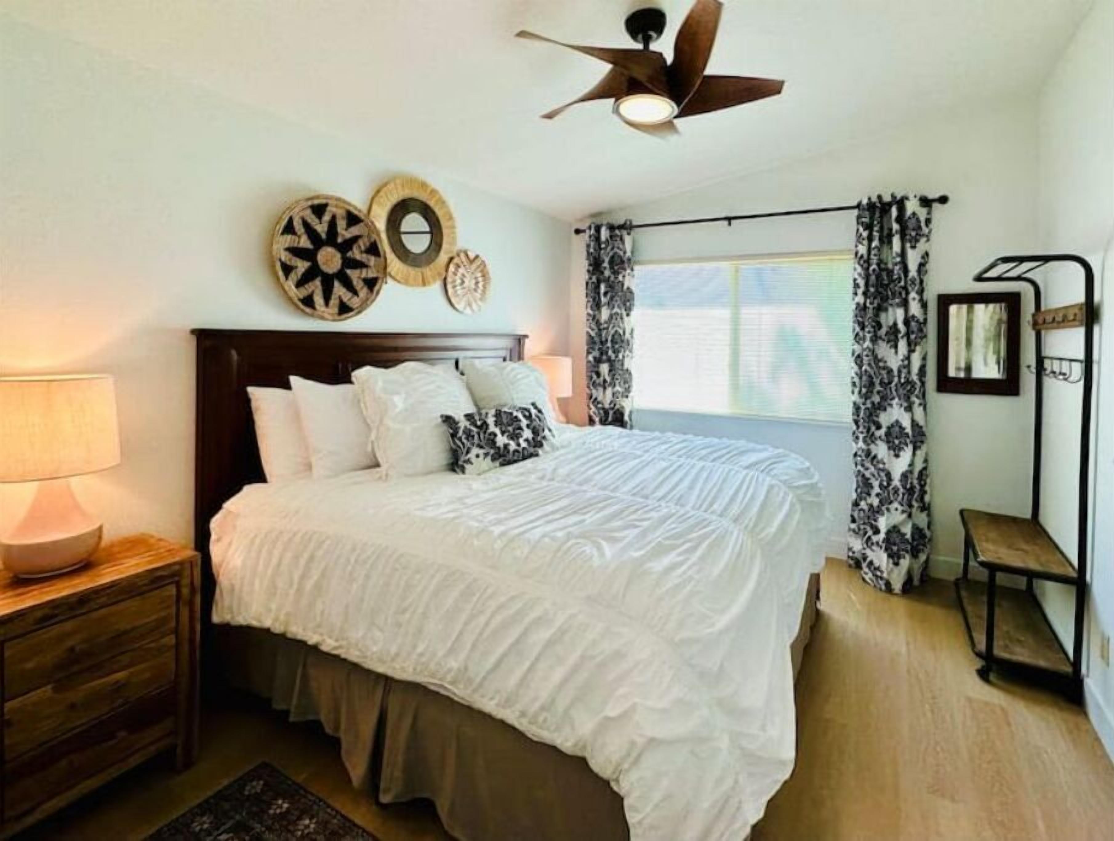 A bedroom with a bed, a nightstand, a wooden dresser, a ceiling fan, and a window with curtains.