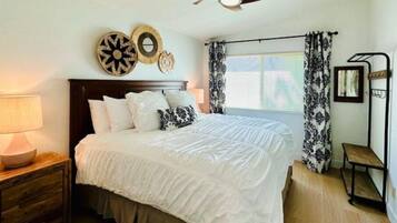 A bedroom with a bed, a nightstand, a wooden dresser, a ceiling fan, and a window with curtains.